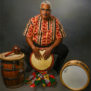 Performer Dimas Sanchez sitting atop a drum with his hands on the drum head